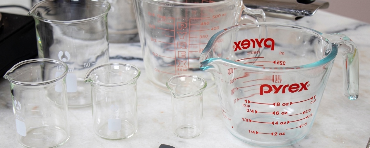 An overhead view of a collection of empty glass laboratory beakers and Pyrex measuring jugs of various sizes arranged on a marble surface.
