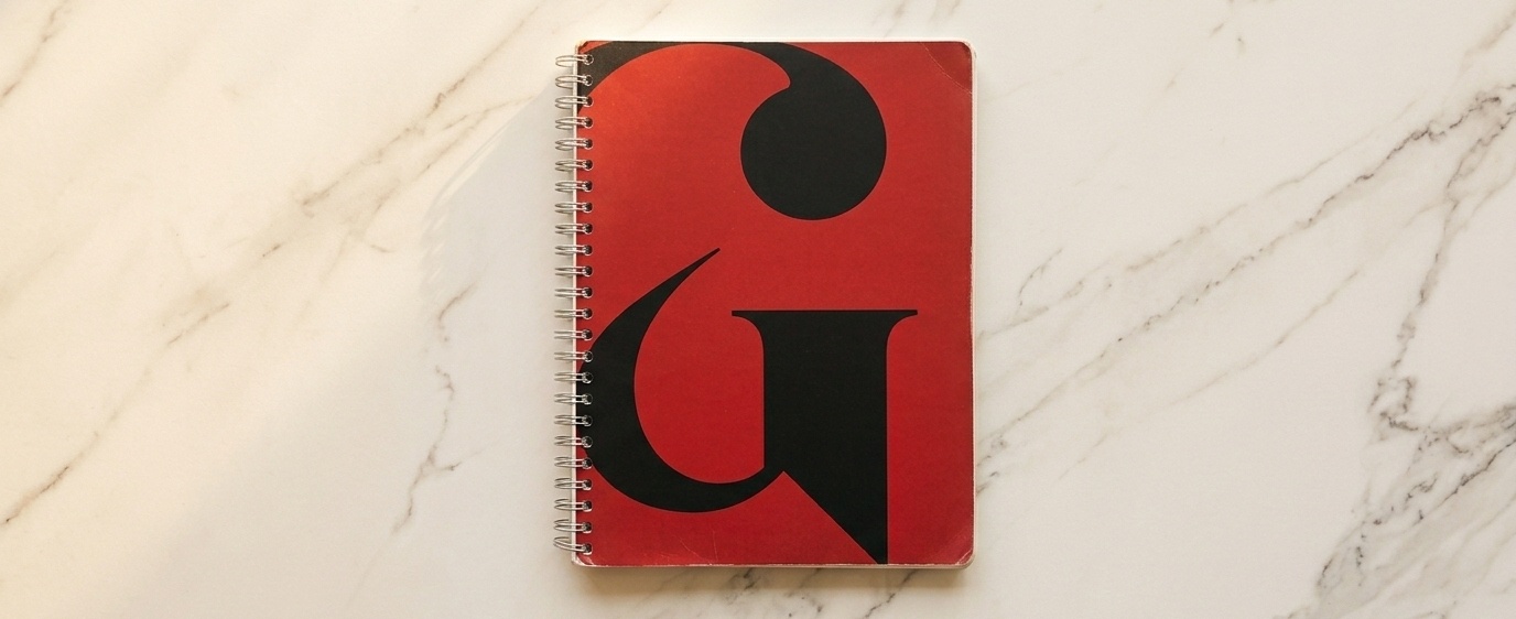 A top-down view of a spiral notebook with a red and black cover, centered on a light-colored marble surface with soft grey veining.
