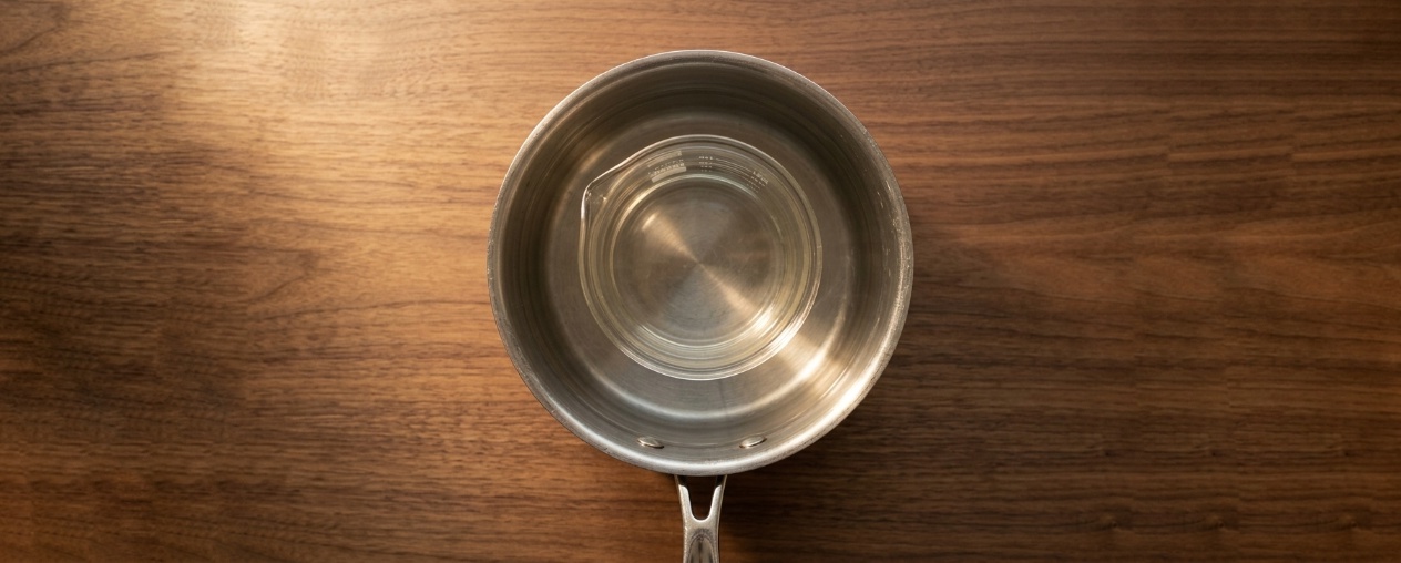A top-down view of a stainless steel pot filled with water, containing a glass beaker, centered on a warm wooden surface.