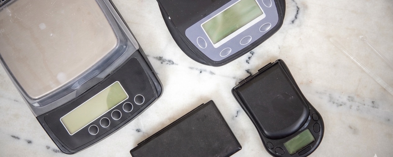 Overhead view of four digital scales on a marble surface: a large top-loading scale, an analytical scale with a transparent chamber, a square scale, and a smaller pocket scale.