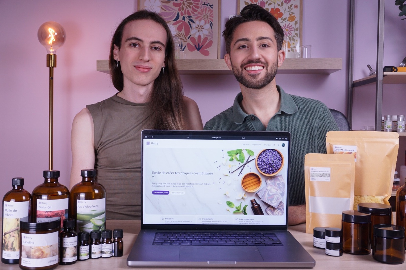 The founders of the Berry DIY smiling behind a laptop displaying the Berry DIY cosmetics website, surrounded by various essential oil bottles and ingredient pouches